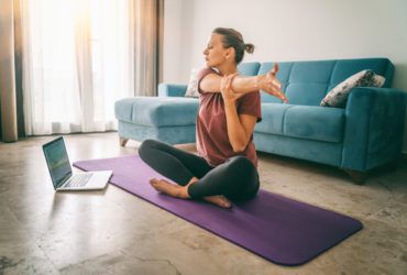 Middle aged woman on yoga mat stretching arm with laptop